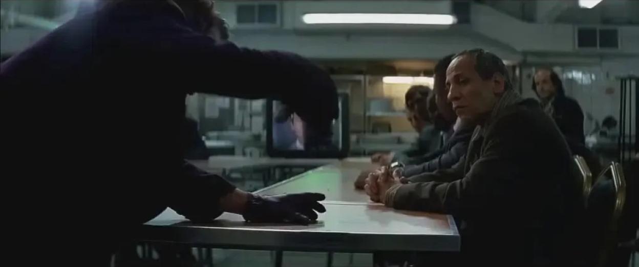 Scene from The Dark Knight: A figure in dark clothing addresses a group of men sitting at a table.