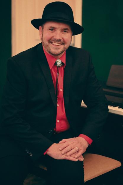 Alan Fletcher, wearing a black suit and hat, sits smiling.