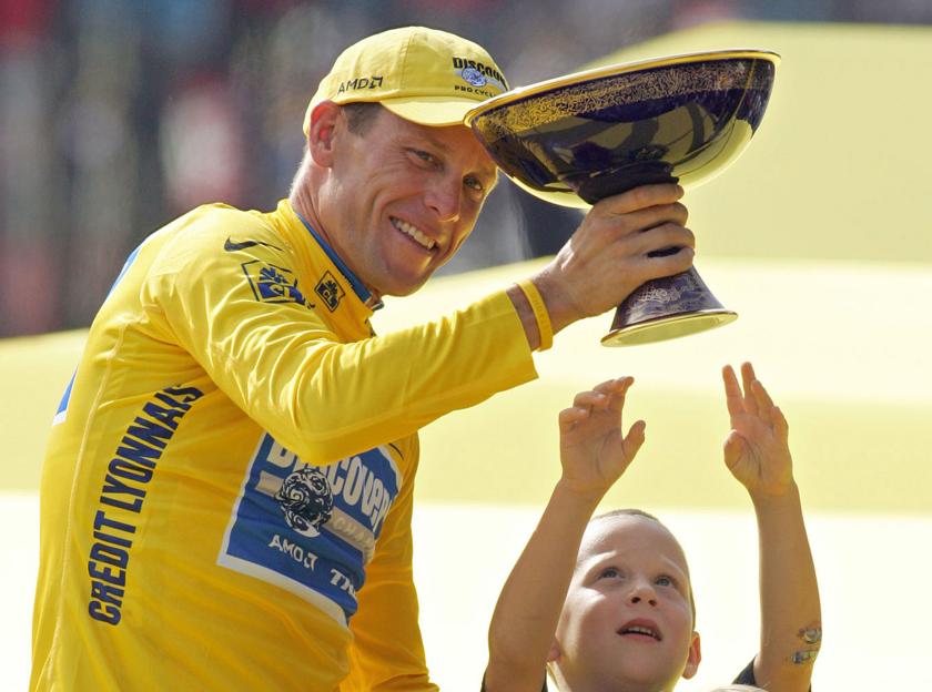Lance Armstrong holding a Tour de France trophy with his son reaching up.