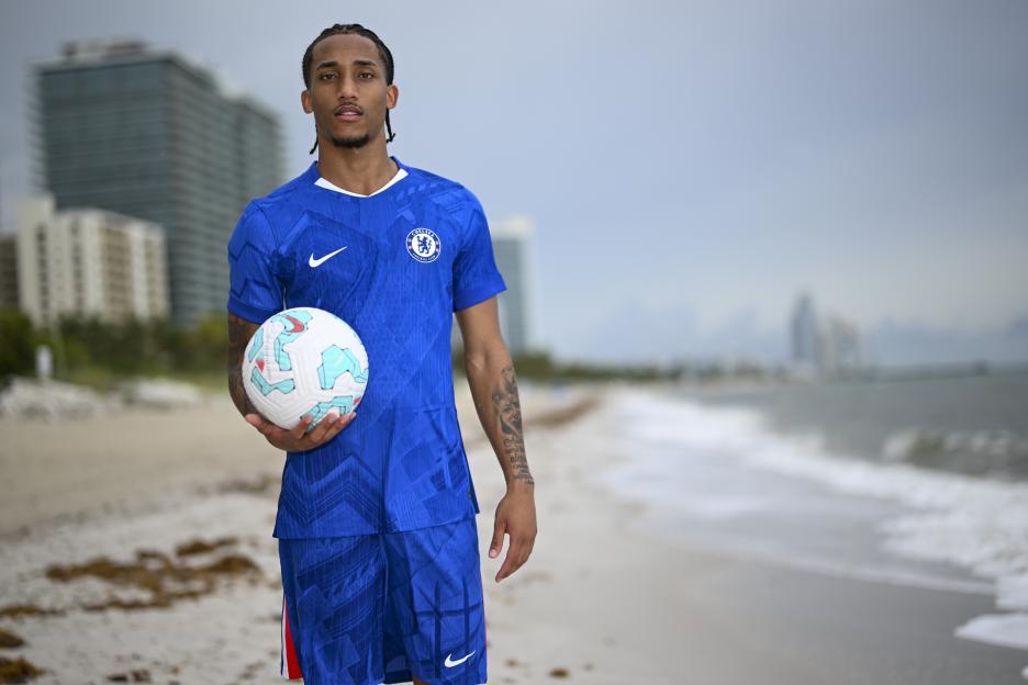 Joao Pedro in Chelsea kit holding soccer ball on beach.