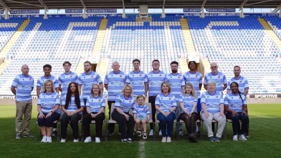 Group photo of people wearing Reading FC's new home kit in front of a stadium.