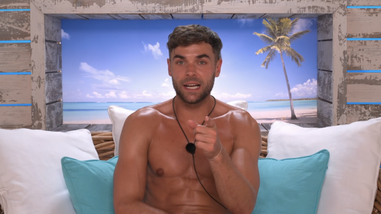 Shirtless man pointing at the viewer, sitting on a couch with a beach scene in the background.