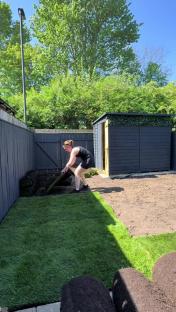 A person laying sod in a backyard next to a dark-colored shed.