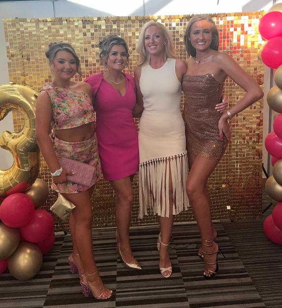 Four women in party dresses pose in front of a gold backdrop.