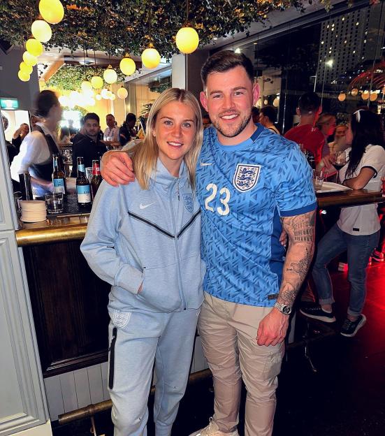 Two people in England athletic wear pose for a photo in a bar.
