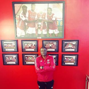 Man in Arsenal jacket standing in front of framed photos of Arsenal teams and players.