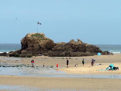 Beach scene with people and a rock formation featuring a Cornish flag.