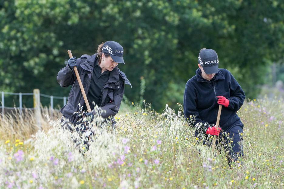 Police officers searching tall grass with shovels at a crime scene.