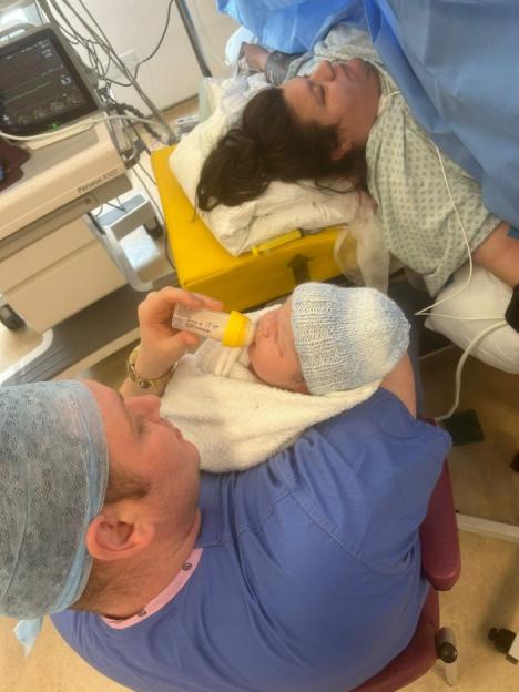 A father feeds his newborn son in a hospital delivery room while the mother rests nearby.