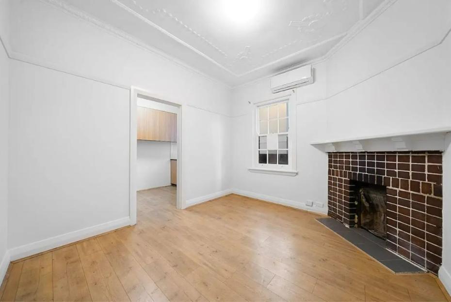 Empty living room with hardwood floors, fireplace, and kitchen view.