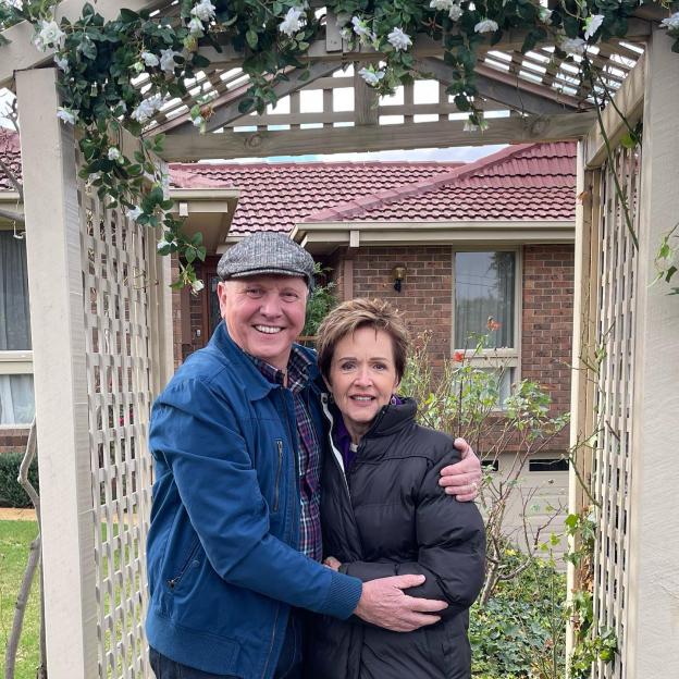 Alan Fletcher and Jackie Woodburne, Neighbours stars, embracing under a garden trellis.