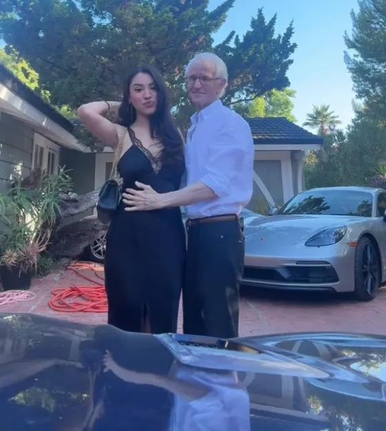 A young woman and an older man stand together in front of a Porsche.