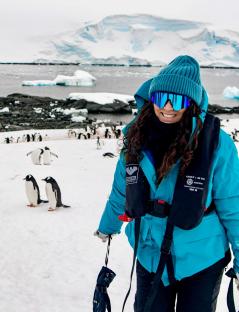 Woman in teal jacket and hat stands in snowy Antarctic landscape with penguins in the background.