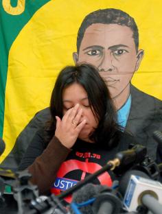 A woman crying at a press conference, with a poster of Jean Charles de Menezes behind her.