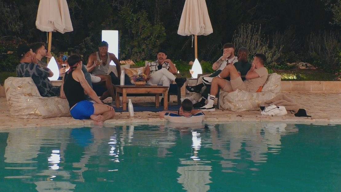 Screengrab of several men sitting by a pool at night, talking.
