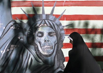 A veiled woman walks past graffiti of a skull-like Statue of Liberty on a wall painted with red and white stripes.