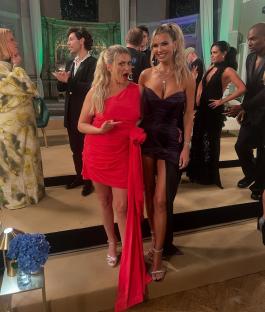 Two women in formal wear pose for a photo with other people in the background.