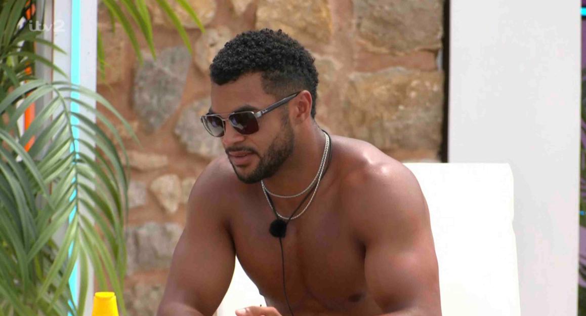 Shirtless man wearing sunglasses sits outdoors.
