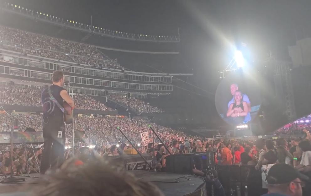 A musician playing guitar on stage in front of a large crowd at a stadium; a large screen behind him shows a couple embracing.
