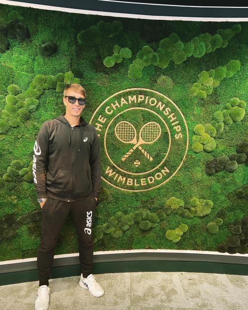Man in Asics tracksuit standing in front of Wimbledon Championships logo on a moss wall.