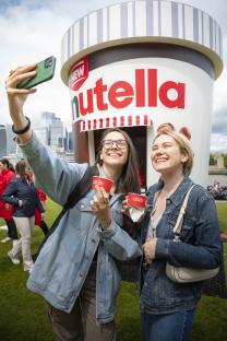 Two friends take a selfie in front of a giant Nutella ice cream tub.