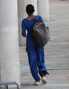 A woman in a blue jumpsuit and gray backpack walks away from the camera.