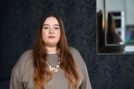 Woman with long auburn hair wearing a taupe sweater and a seashell necklace.