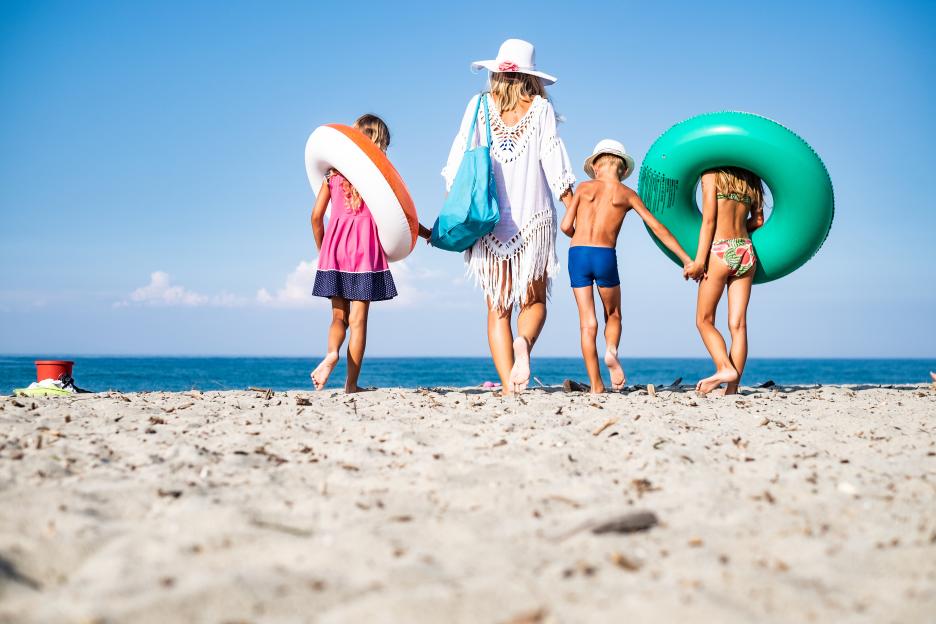 Rear view of a woman and three children walking on a beach, carrying inflatable rings.