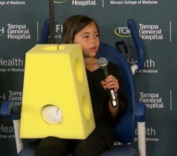 Girl holding microphone, wearing a yellow foam block costume.