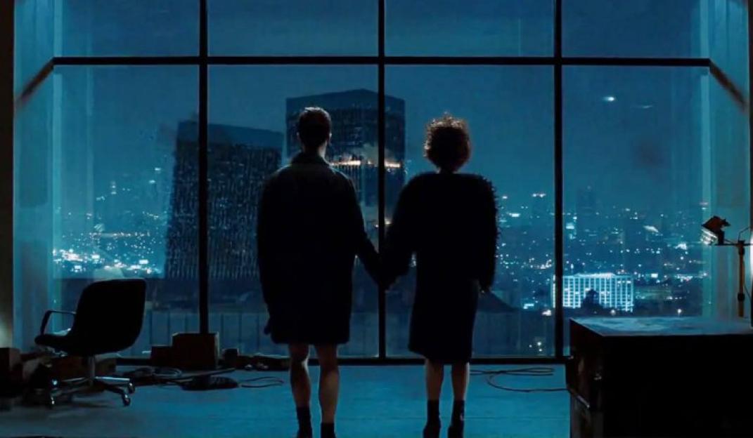 Film still of two people holding hands in front of a large window overlooking a city at night.