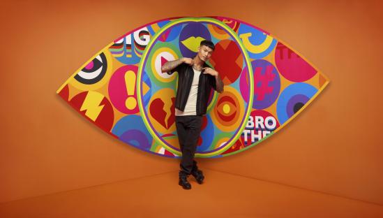 A Big Brother contestant posing in front of a colorful eye-shaped graphic.