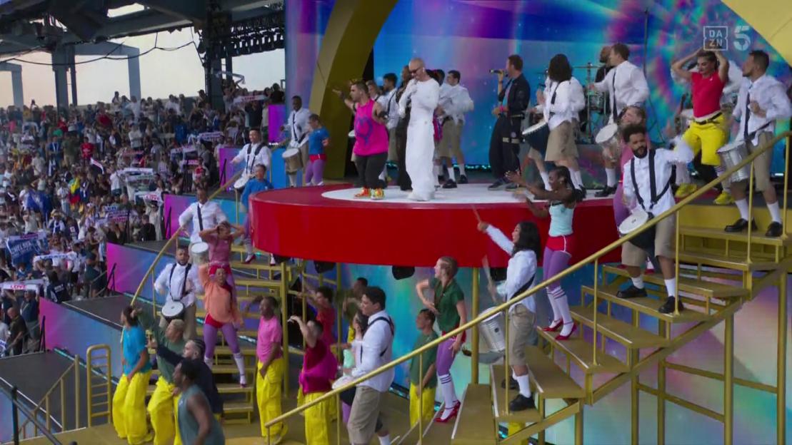 Large group of performers playing drums on a stage in front of an audience.
