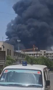 Large plume of black smoke billowing over buildings, ambulance in foreground.