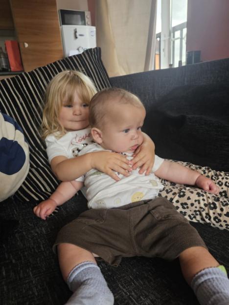 A toddler girl hugging a large baby boy on a couch.