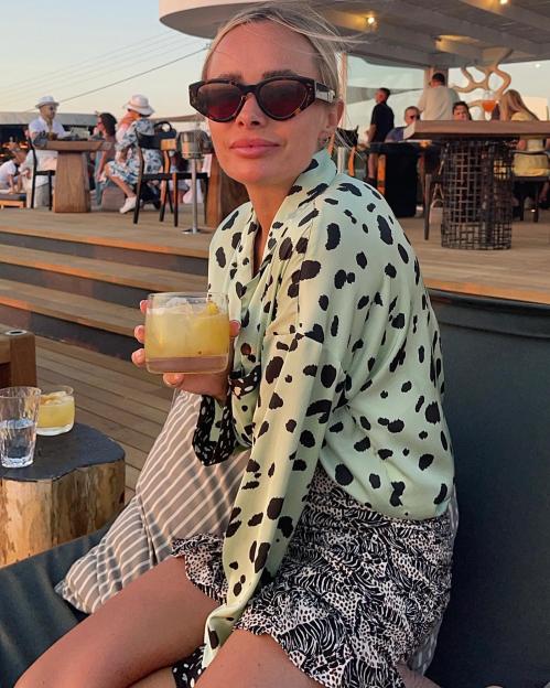 Woman in patterned shirt and skirt holding a cocktail on a patio.
