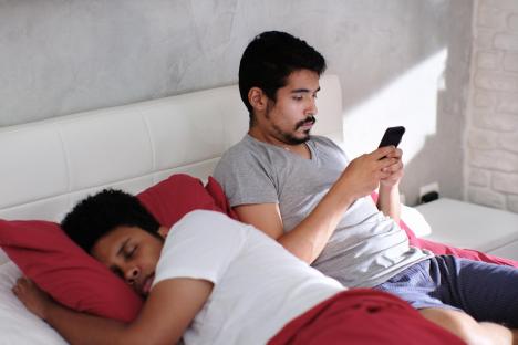 A man secretly texting on his phone while his partner sleeps beside him in bed.