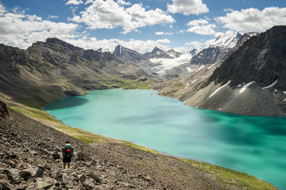 A hiker pauses on a rocky trail overlooking a turquoise lake and snow-capped mountains in Kyrgyzstan.