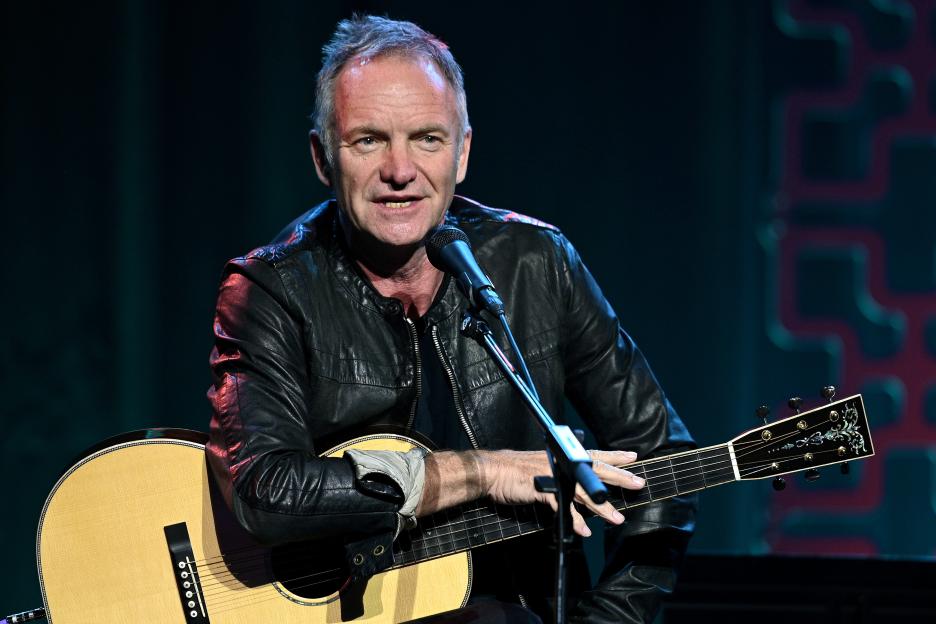 Sting performing live on stage with an acoustic guitar.