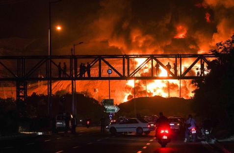 People watch a large fire at an oil refinery at night.