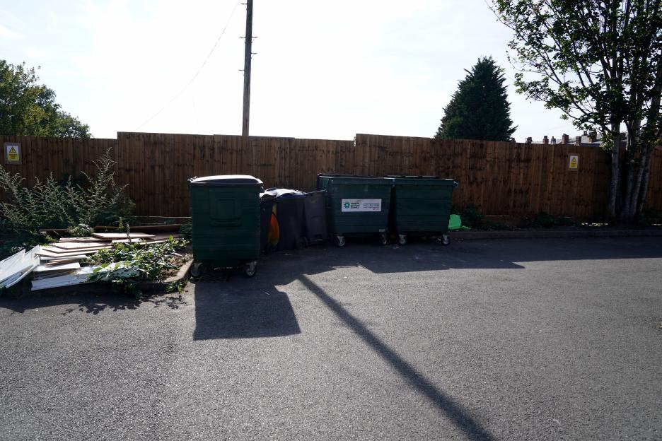 Green wheelie bins and discarded building materials outside a wooden fence.