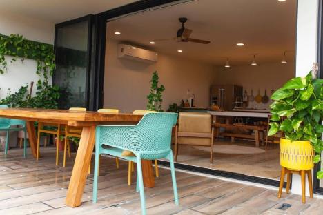 Outdoor patio with wooden table and chairs, overlooking a brightly lit interior kitchen and living space.