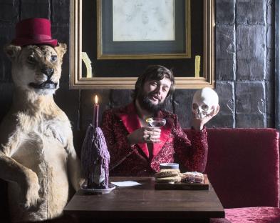 A man in a red sequined jacket sits at a table with a taxidermied lion wearing a top hat, a skull, and a cocktail.