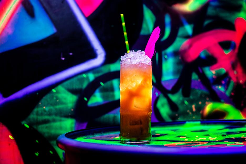 Cocktail in tall glass with ice and straws in front of a neon graffiti wall.