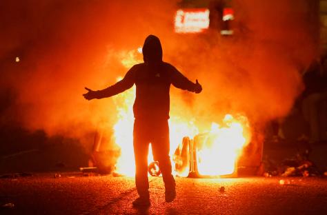 A person silhouetted against a burning vehicle during a riot.