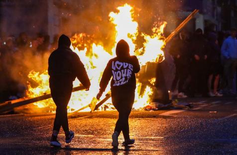 Two people walk away from a large fire during a nighttime protest.