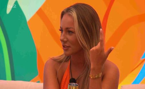 Screenshot of a blonde woman wearing an orange top, making a hand gesture.