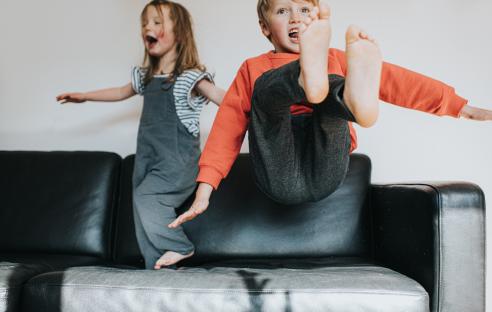 Two children playfully jumping on a black leather sofa.