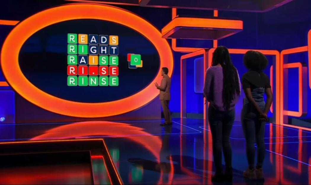 Game show set with word puzzle on screen and contestants.