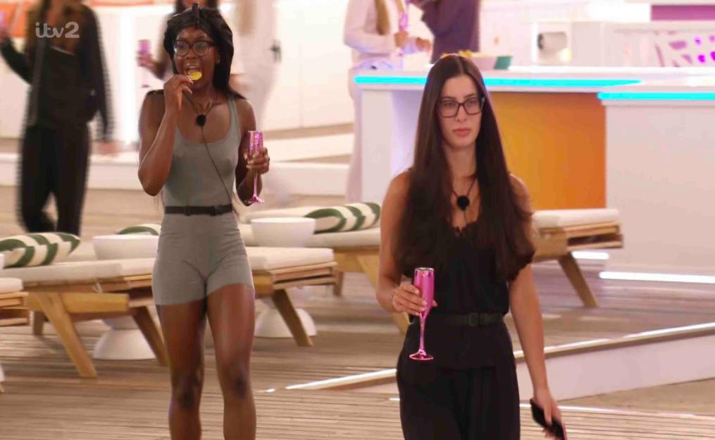 Two women walking, one eating a chip and holding a glass, the other holding a glass.