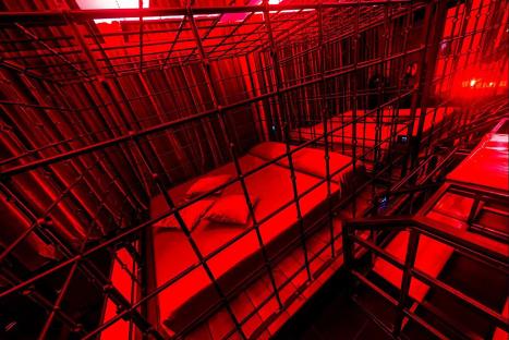 Hotel room beds enclosed in red-lit cages.